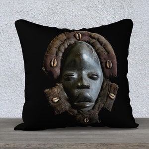 May include: Black and brown throw pillow with a carved wooden mask design. The mask has a dark brown face with a wide mouth and eyes. The mask is surrounded by a brown and red fabric with white shell accents.