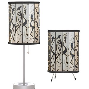 May include: Two table lamps with black and gold patterned lampshades featuring stylized figures. One lamp has a silver base and a pull chain, the other has a tripod base.