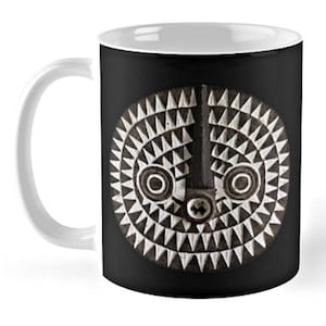 Puede incluir: Una taza de café de cerámica blanca con asa negra. La taza presenta un gráfico en blanco y negro de una máscara tribal con un diseño geométrico.