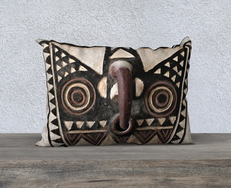 May include: A decorative pillow with a black and white tribal mask design. The mask has two large eyes with concentric circles and a prominent nose with a ring.
