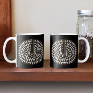 May include: Two white ceramic mugs with black handles. Each mug features a black and white geometric design of a stylized face with a central eye and two smaller eyes on either side.