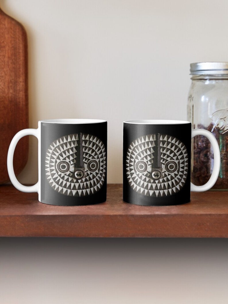 May include: Two white ceramic mugs with black handles. Each mug features a black and white geometric design of a stylized face with a central eye and two smaller eyes on either side.