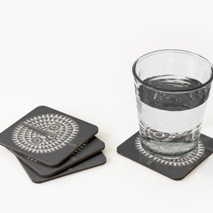 May include: Set of four gray coasters with a white geometric design. The design features a stylized face with two eyes and a mouth.