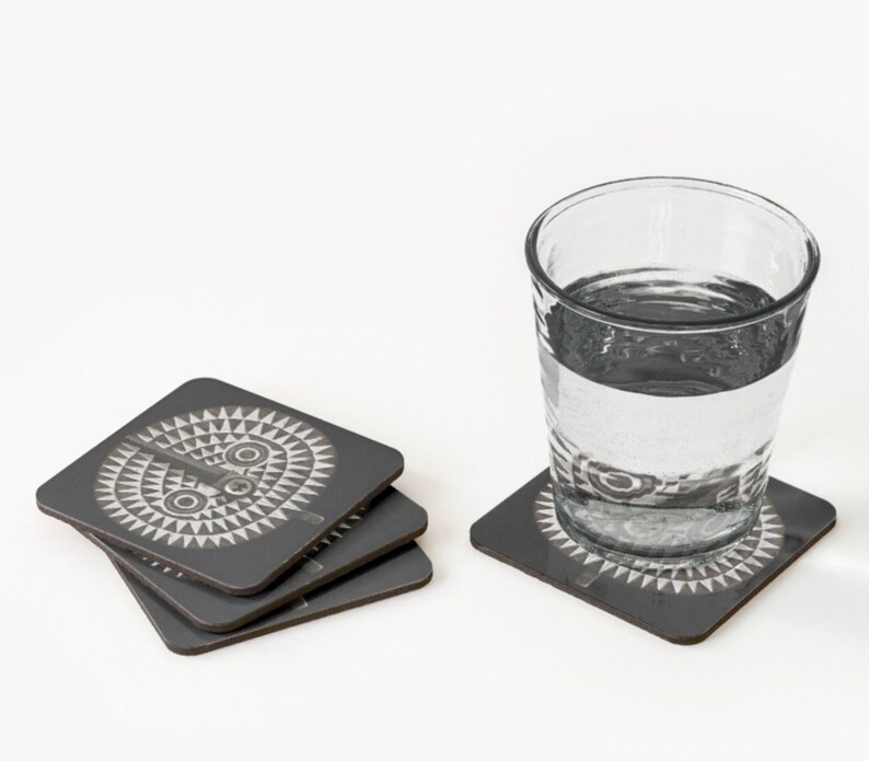 May include: Set of four gray coasters with a white geometric design. The design features a stylized face with two eyes and a mouth.