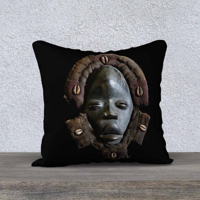 May include: Black and brown throw pillow with a carved wooden mask design. The mask has a dark brown face with a wide mouth and eyes. The mask is surrounded by a brown and red fabric with white shell accents.