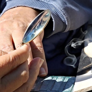 Może przedstawiać: Osoba używa lupy, aby skupić światło słoneczne na kawałku drewna, tworząc dym. Drewno jest spalane, aby stworzyć wz&oacute;r.