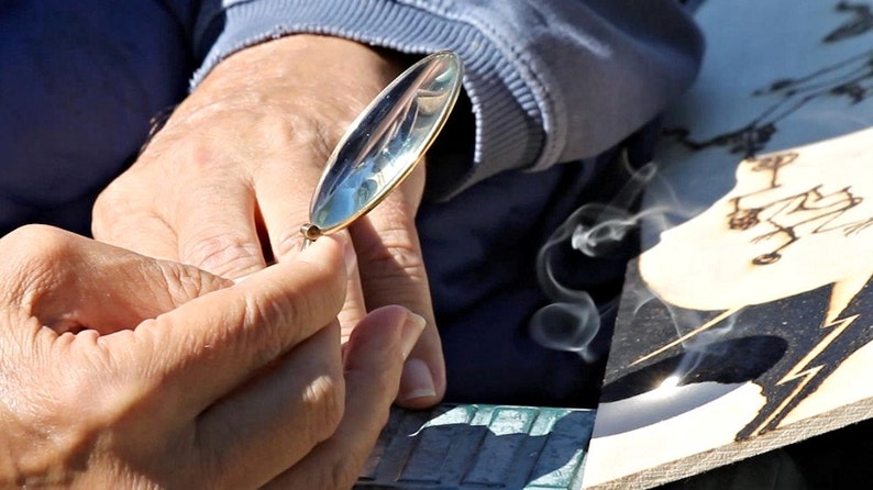 Może przedstawiać: Osoba używa lupy, aby skupić światło słoneczne na kawałku drewna, tworząc dym. Drewno jest spalane, aby stworzyć wz&oacute;r.