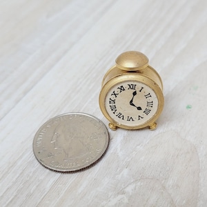 May include: A small gold-colored alarm clock with Roman numeral markings on the face. The clock is on a light wood surface.
