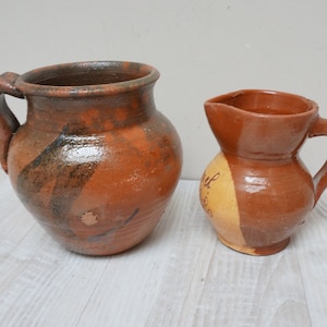 May include: Two terracotta pottery pitchers, one larger than the other. The larger pitcher has a brown and green glaze, while the smaller pitcher has a brown and yellow glaze.