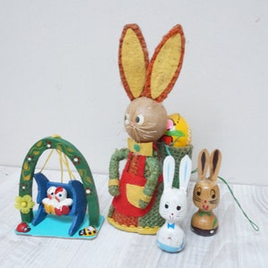 May include: Three wooden Easter bunny figurines. The largest bunny is wearing a red and green dress and carrying a basket with a yellow egg. The other two bunnies are smaller and are white and brown.