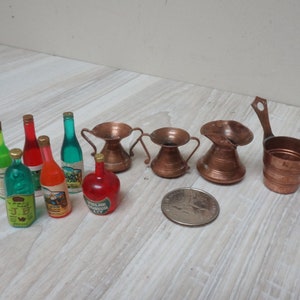 May include: A set of miniature copper kitchenware and glass bottles. The set includes three copper pitchers, a copper measuring cup, and six glass bottles with labels. The bottles are filled with green, red, and clear liquids.