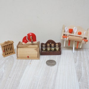 May include: Miniature wooden kitchen accessories, including a bread box with a red handle, a spice rack with three glass jars, and a wall-mounted utensil holder with a rolling pin. The set also includes a small wooden cabinet.