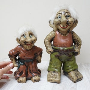 May include: Two ceramic troll figurines with white hair. The larger troll is wearing a red tank top and green trousers. The smaller troll is wearing a brown robe. Both trolls have a whimsical expression.