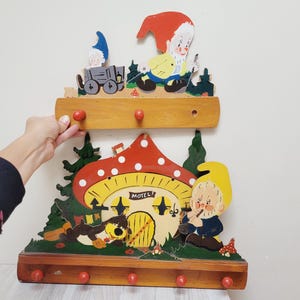 以下が含まれることがあります： 塗装されたノームのイラストが描かれた2段の木製ウォールラック。上段には3つの赤いフックと赤い帽子のノーム、下段には5つの赤いフック、MOTELと書かれたキノコ型の家、笛を吹くノームが描かれています。