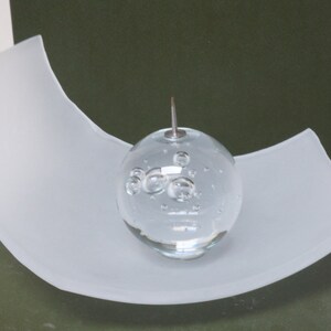 May include: A clear glass sphere with air bubbles inside, resting on a white curved glass base.