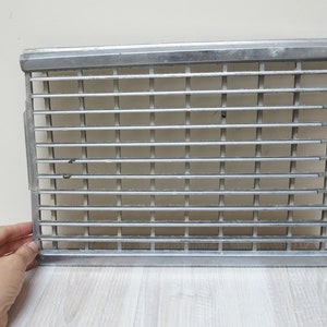 Puede incluir: Una rejilla de metal plateado con forma rectangular y una serie de barras horizontales. La rejilla tiene una superficie ligeramente texturizada y parece estar hecha de un material duradero.