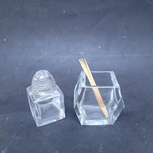 Può includere: Due contenitori di vetro trasparente. Uno è una piccola bottiglia quadrata con un coperchio a cupola strutturato. L'altro è un contenitore sfaccettato a forma esagonale con alcuni stuzzicadenti di legno all'interno. Entrambi sono su uno sfondo scuro.