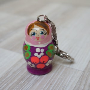 Op de afbeelding: Een paarse en roze houten Matroesjka-sleutelhanger met een geschilderd gezicht en een bloemenpatroon. De sleutelhanger heeft een zilveren ketting.