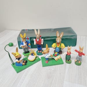 Può includere: Un set di sette figurine di coniglietti pasquali in legno. I coniglietti sono dipinti in vari colori, tra cui giallo, blu, rosso, verde e bianco. Indossano abiti diversi e tengono in mano oggetti diversi, come un cestino, uno strumento musicale e un carretto. Le figurine sono disposte su una superficie di legno.