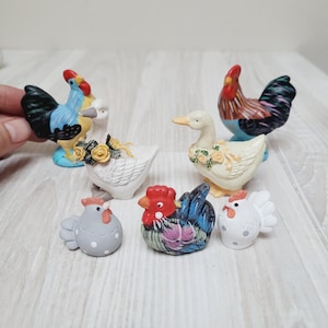 May include: A selection of ceramic chicken and duck figurines. The figurines are painted in various colours, including blue, red, white, and yellow. Some are decorated with floral accents. The collection includes roosters, hens, and ducks.