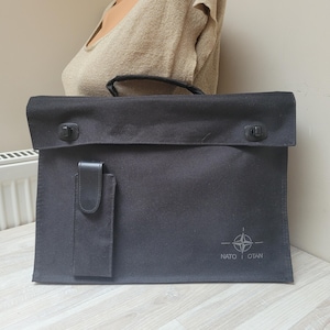 May include: A black rectangular briefcase made of durable fabric featuring a top handle, a fold-over flap with two black plastic fasteners, a vertical strap on the front, and the NATO OTAN emblem printed in the bottom right corner.