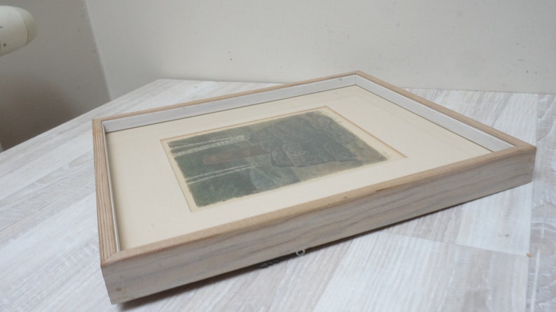 May include: A framed print of a landscape scene with trees and a building in the distance. The print is in shades of gray and brown and is set against a white background.