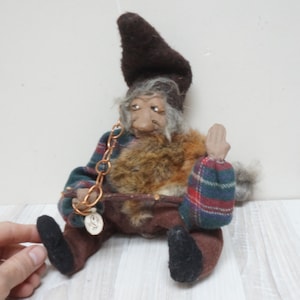 Könnte beinhalten: Eine Plüsch-Gnom-Puppe mit einem braunen Hut, einem karierten Hemd und einer braunen Hose. Der Gnom hat einen langen weißen Bart und hält eine kleine Kette mit einem runden Metalletikett.