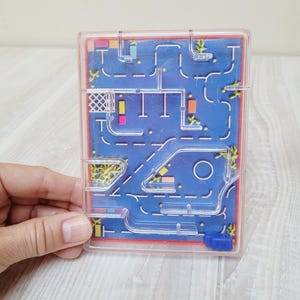 May include: A clear plastic maze game with a blue background featuring a road map design. The maze has a series of interconnected pathways and obstacles. The game is held in a person's hand.
