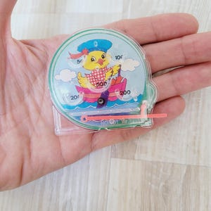 Könnte beinhalten: Ein durchsichtiges, tragbares Wasserspiel aus Kunststoff mit einer gelben Ente mit Matrosenmütze in einem rosa Boot. Das Spiel hat einen blauen Hintergrund mit weißen Wolken und Zahlen am Rand. Der Text "MADE IN CHINA" ist sichtbar.