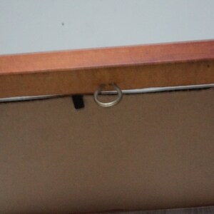 May include: Close-up of the back of a framed artwork. The frame is made of wood with a reddish-brown finish. A metal ring is attached to the frame for hanging. The artwork itself is a light brown color.