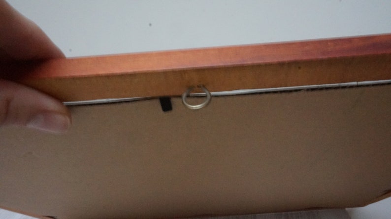 May include: Close-up of the back of a framed artwork. The frame is made of wood with a reddish-brown finish. A metal ring is attached to the frame for hanging. The artwork itself is a light brown color.