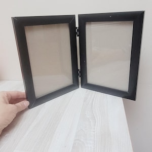 Könnte beinhalten: Ein schwarzer Doppelbilderrahmen mit zwei leeren Öffnungen. Der Rahmen ist geöffnet und liegt auf einer hellbraunen Holzoberfläche.