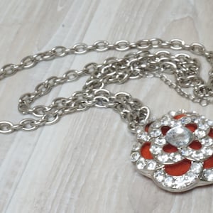 May include: A silver chain necklace with a large, red and silver flower pendant. The flower is adorned with clear rhinestones.