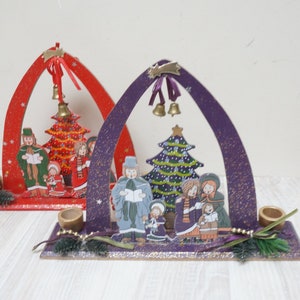 K&ouml;nnte beinhalten: Zwei h&ouml;lzerne Weihnachtsb&ouml;gen mit Kerzenhaltern. Die B&ouml;gen sind mit gemalten Szenen von Familien verziert, die Weihnachtslieder um einen Weihnachtsbaum singen. Ein Bogen ist rot und der andere ist lila.