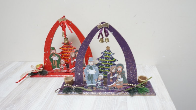K&ouml;nnte beinhalten: Zwei h&ouml;lzerne Weihnachtsb&ouml;gen mit Kerzenhaltern. Die B&ouml;gen sind mit gemalten Szenen von Familien verziert, die Weihnachtslieder um einen Weihnachtsbaum singen. Ein Bogen ist rot und der andere ist lila.