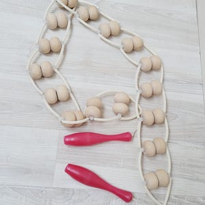 May include: A wooden massage roller with two red handles. The roller features multiple pairs of wooden balls connected by a white flexible cord. The handles are a bright, solid red color.