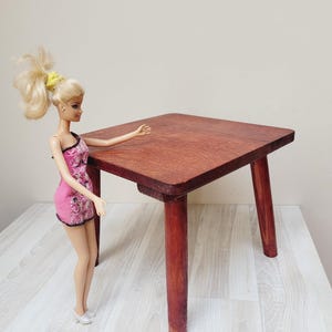 Puede incluir: Una pequeña mesa cuadrada de madera con cuatro patas. La mesa está teñida de color marrón rojizo. Una muñeca con un vestido rosa está de pie junto a la mesa.