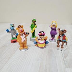 May include: A collection of seven Muppet Show character figurines. The figurines include Kermit the Frog, Miss Piggy, Fozzie Bear, Gonzo, and others. The figures are brightly colored and stand on small, round bases.