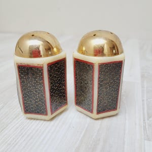 Op de afbeelding: Een paar vintage zout- en peperstrooiers. Elke strooier heeft een goudkleurige, afgeronde bovenkant met kleine gaatjes. Het onderste deel is crèmekleurig met een zwart en rood patroon. De strooiers staan op een licht houten oppervlak.