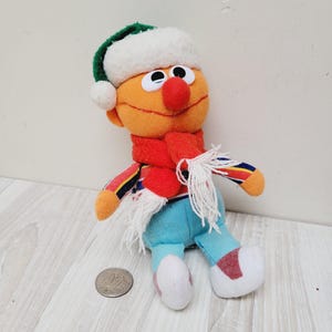Könnte beinhalten: Ein orangefarbener Plüschcharakter aus der Sesamstraße, der eine weiße und grüne Weihnachtsmütze, einen roten Schal und eine blaue Hose trägt. Der Charakter sitzt auf einer hellbraunen Holzoberfläche.