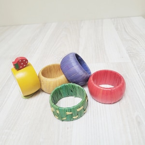 Könnte beinhalten: Fünf bunte gewebte Serviettenringe. Die Ringe sind aus natürlichen Materialien gefertigt und haben einen rustikalen Look. Ein Ring hat eine kleine Holzerdbeere daran befestigt.