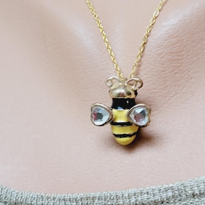 May include: A gold-colored necklace with a bee pendant. The bee pendant is black and yellow with clear, heart-shaped wings. The necklace is worn on a person.