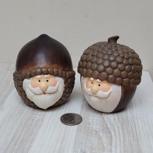 May include: Two ceramic acorn figurines with Santa Claus faces. The acorns are brown with white fur trim and white faces with black eyes, red cheeks, white beards, and black eyebrows.