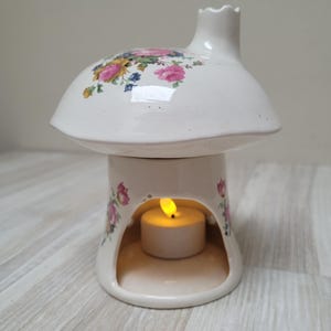 Pode incluir: Um queimador de óleo em cerâmica branca em forma de cogumelo com detalhes florais. A parte superior apresenta flores rosa, amarelas e azuis. Uma vela de chá acesa está dentro da base, visível através de uma abertura arqueada.