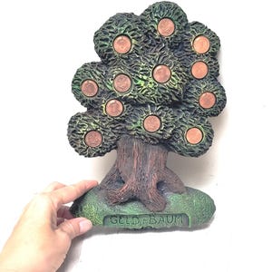 Puede incluir: Un árbol de dinero de cerámica verde y marrón con 10 ranuras para monedas. La base del árbol tiene la inscripción "GELD-BAUM".