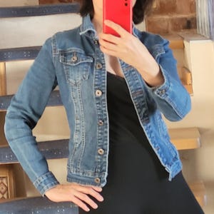 Peut inclure: Veste en jean bleu moyen, avec des boutons et des poches. La veste est portée avec un haut et un legging noirs. L'image est prise à l'intérieur, avec un escalier en arrière-plan.