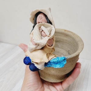 May include: A handmade ceramic planter featuring a seated figure in a hat, holding a baby wrapped in a blanket. The planter has a beige and blue colour scheme, with a textured finish. The figure is seated on the edge of the planter.