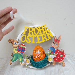 Puede incluir: Una corona circular blanca con una pancarta amarilla que dice "Frohe Ostern" y dos conejos que sostienen huevos de Pascua. La corona está decorada con flores blancas y un nido con un huevo de Pascua decorado.