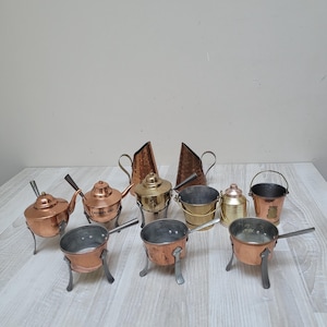 Puede incluir: Una colección de miniaturas de utensilios de cocina de cobre y latón, que incluyen teteras, ollas, cubos y jarras. Los artículos están dispuestos sobre una superficie de madera clara, mostrando sus diversas formas y acabados metálicos.
