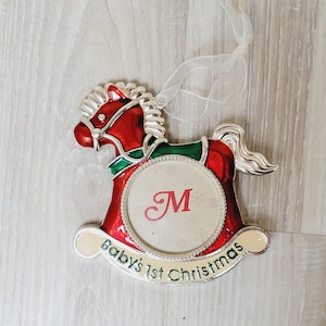 Puede incluir: Adorno de Navidad de caballo de balancín rojo y verde con un marco plateado y un fondo blanco. El marco tiene una letra "M" en el centro. El adorno tiene el texto "Baby's 1st Christmas" en la parte inferior.